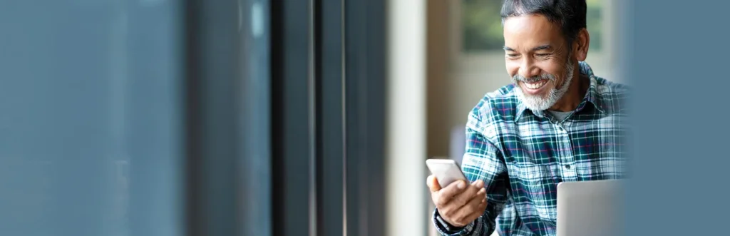 A man in a plaid shirt smiles while using a smartphone, seated next to a laptop in a bright indoor setting.