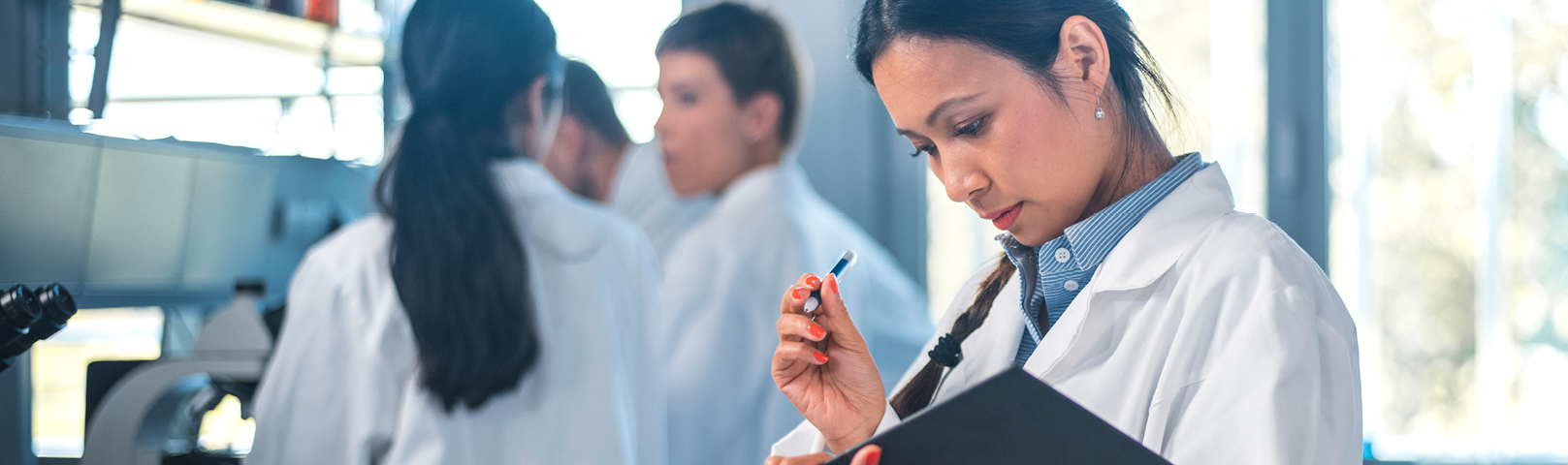 a latino woman in a lab coat reviews her clip board carefully just next to a microscope in a lab